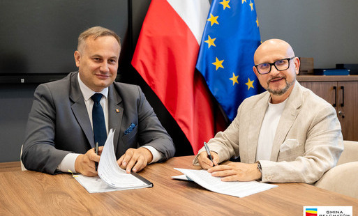 wicemarszałek piotr wojtysiak i wójt konrad koc siedzą przy stole i podpisują dokumenty. W tle flaga polska i flaga unijna